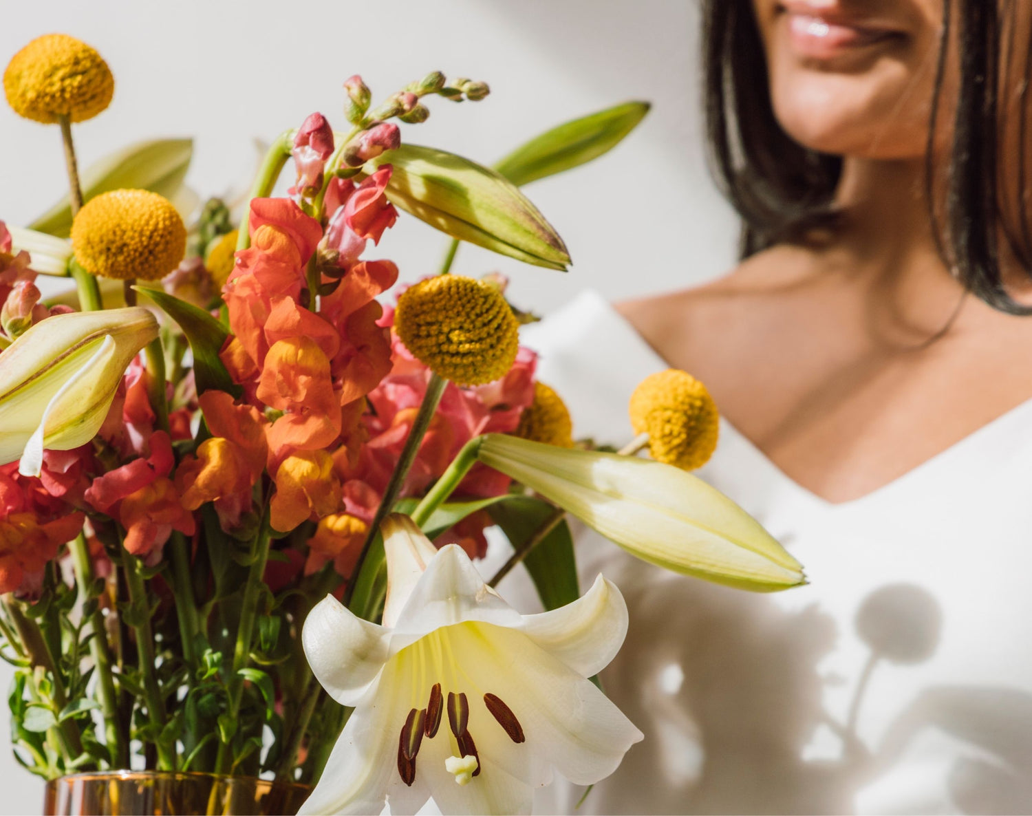 A close-up of a vibrant flower bouquet with white lilies, orange snapdragons, and yellow craspedia, styled next to a woman in a white top.
